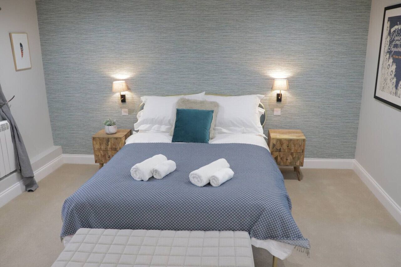 A neatly made bed with two pillows, a blue blanket, and two rolled towels on the bed. There are two bedside tables with lamps, a small plant, and a framed picture on the wall.