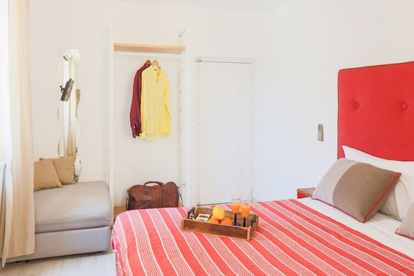 Un dormitorio con cama, mesita de noche, perchero, maleta y bandeja de madera con frutas y jugo.
