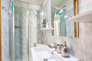 Un baño moderno con ducha envuelta en vidrio, un lavabo blanco con espejo y un estante con artículos de aseo.