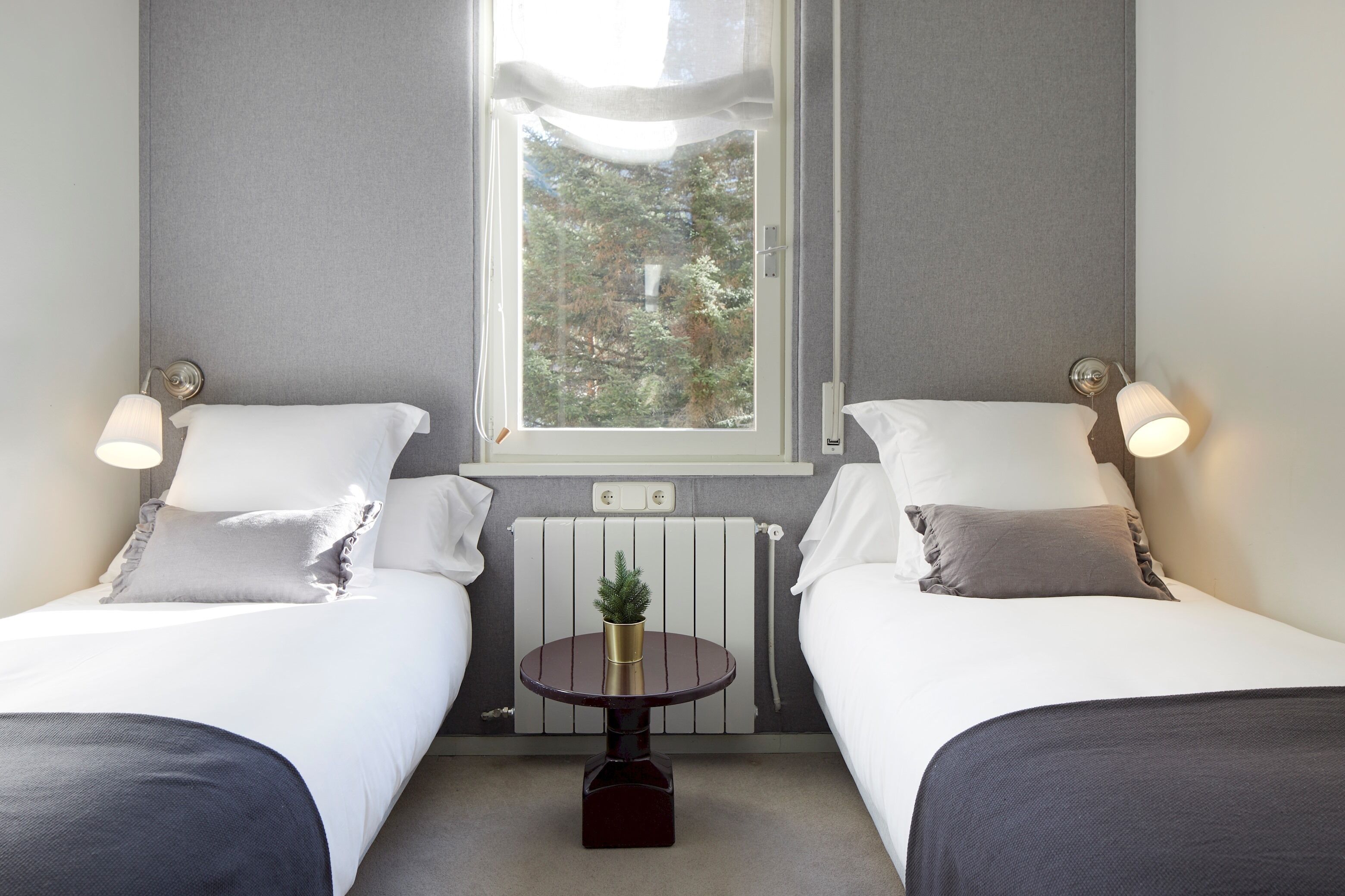 A hotel room with two beds, a small round table with a vase, and a window with a view of trees.