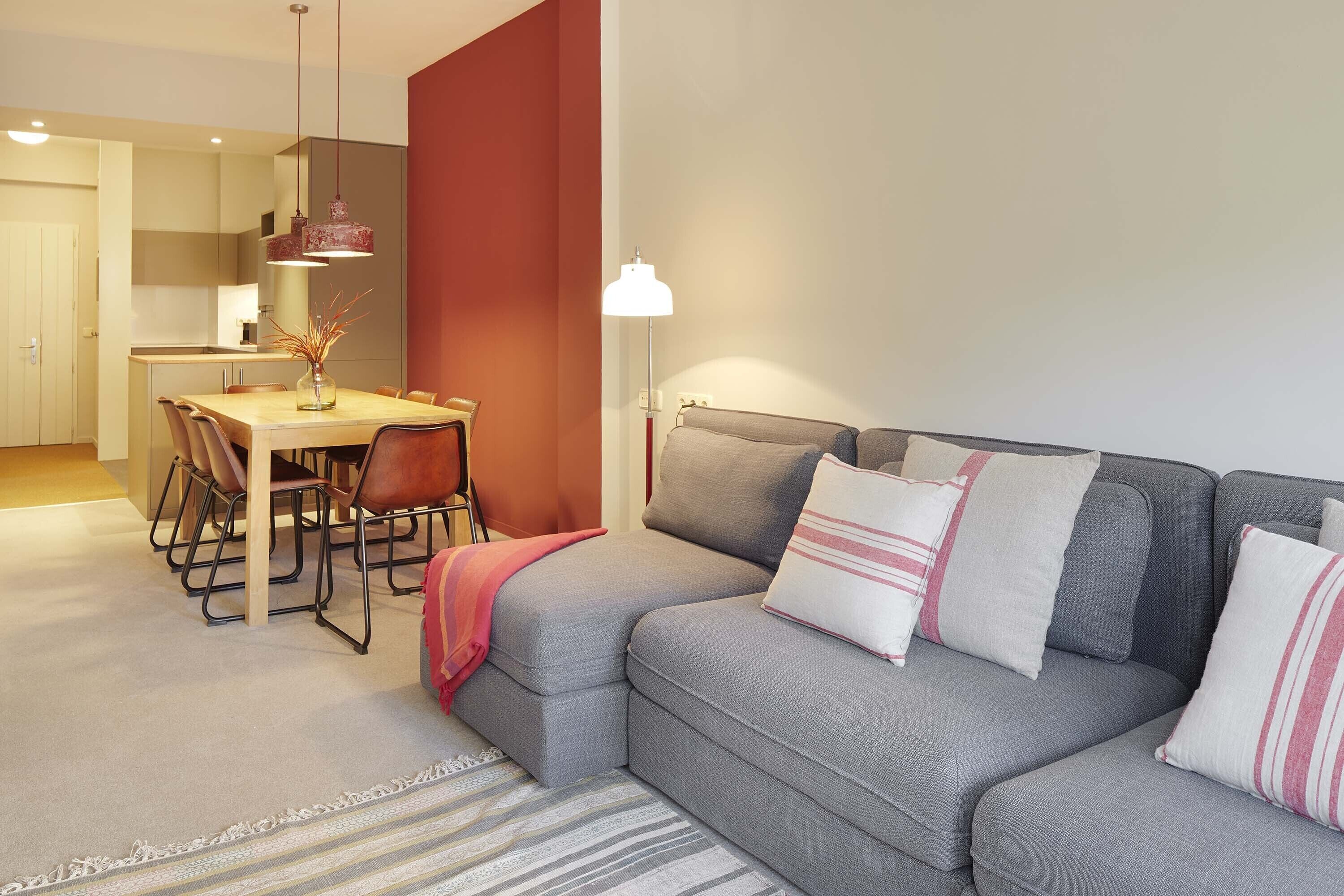 A modern living room with a grey sofa, a dining table with chairs, and a kitchen in the background.