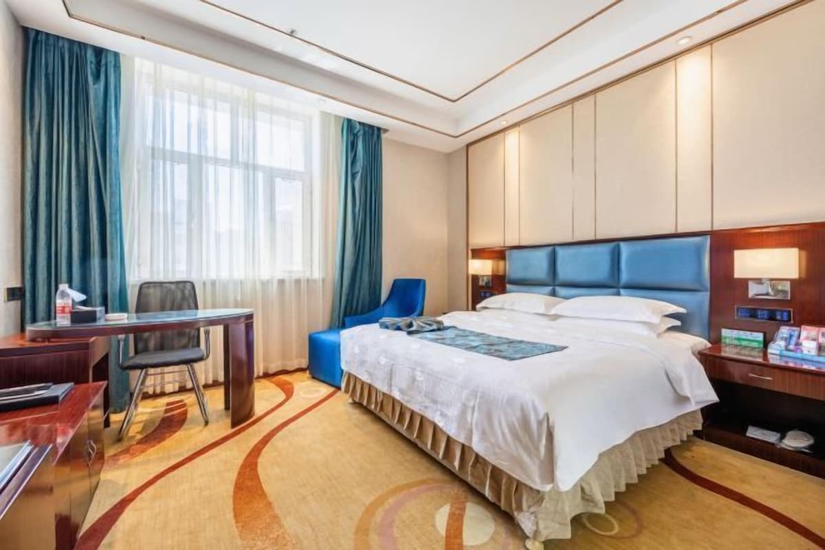 Habitación de hotel con una cama grande, un escritorio, una silla y un ventanal con cortinas.