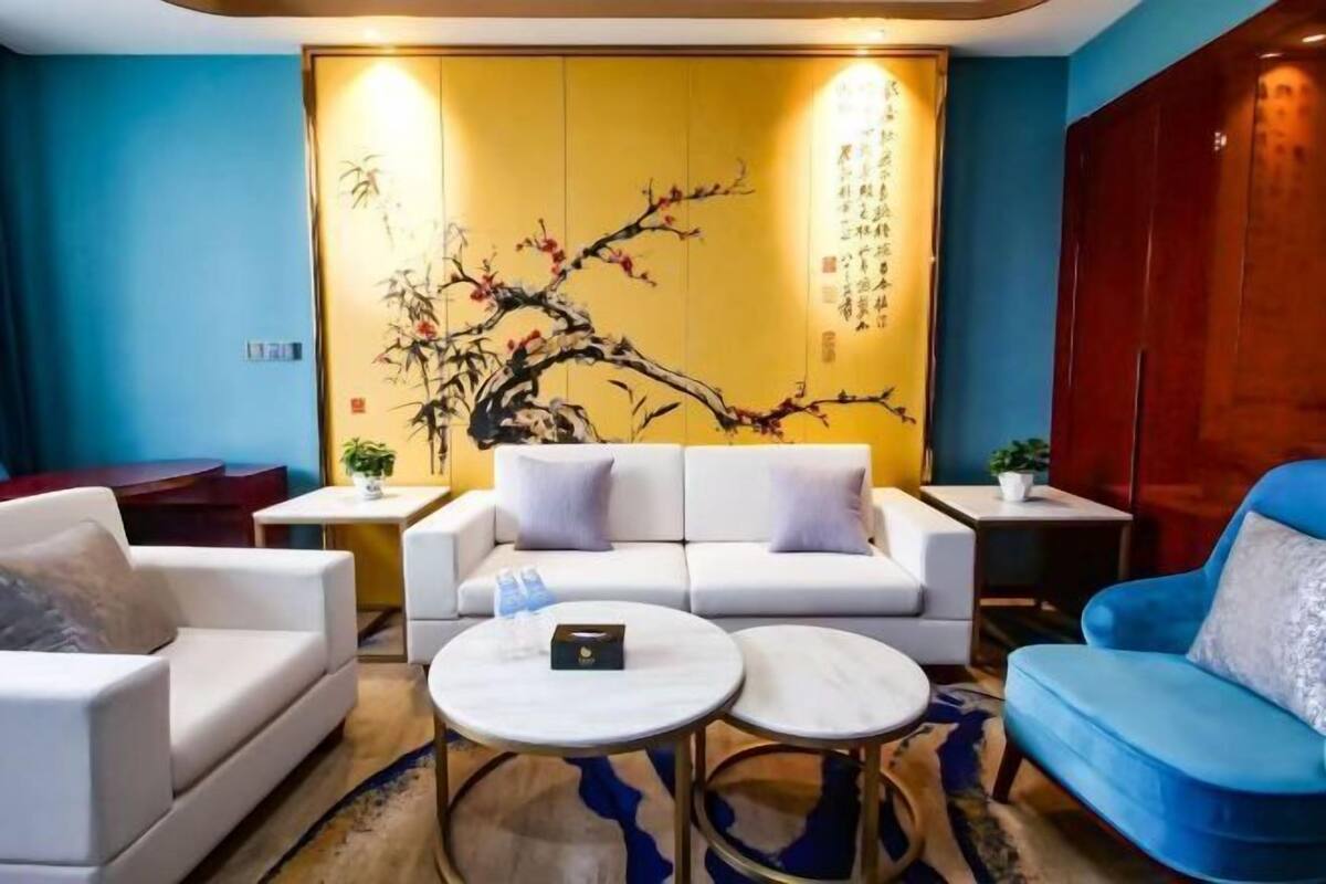 A modern living room with a white sofa, blue armchairs, and a round coffee table. A large wall mural with Chinese calligraphy and a tree is visible.