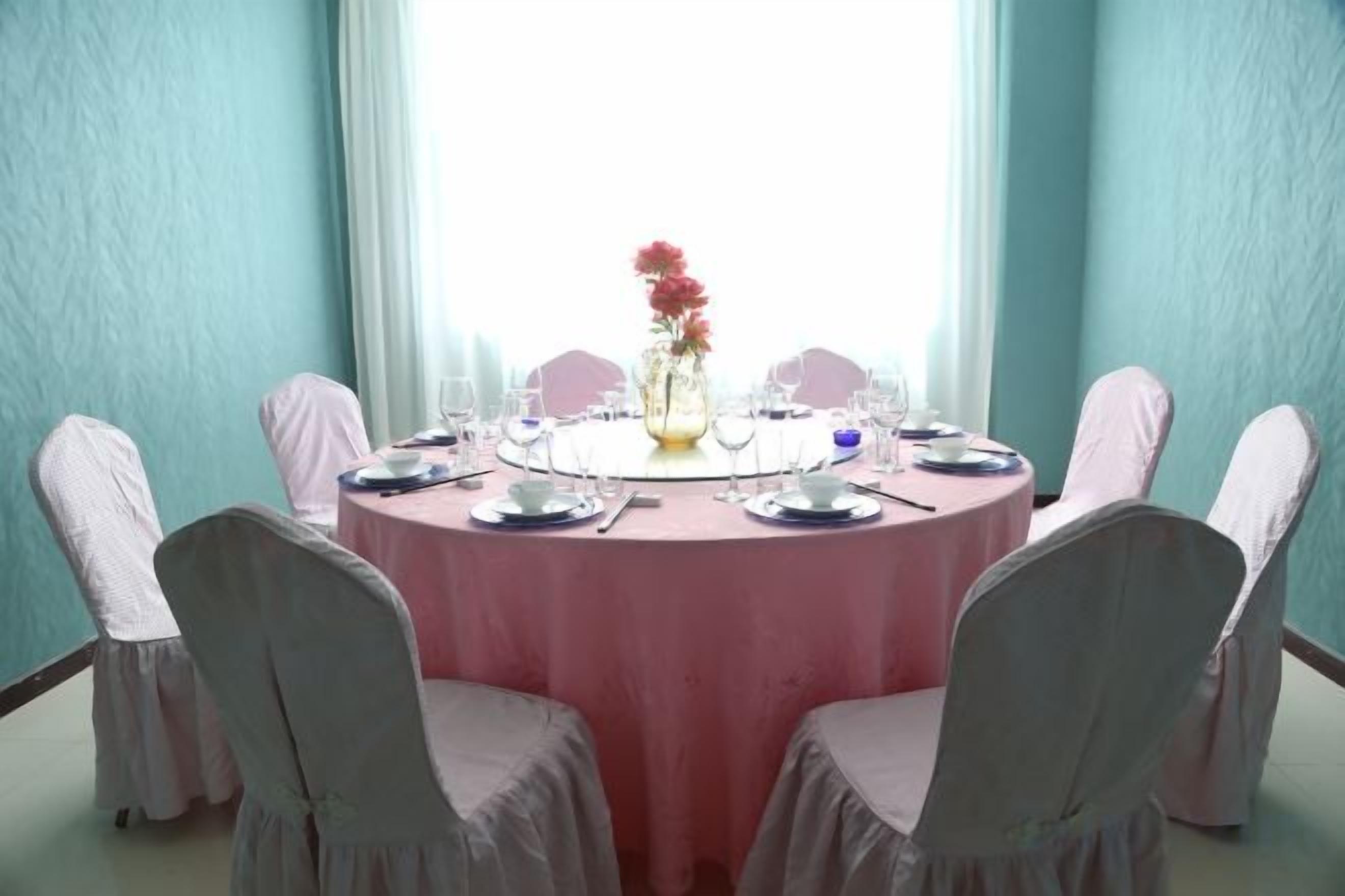 A round table with a pink tablecloth, surrounded by chairs with white covers, set for a meal.