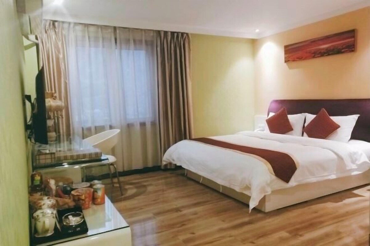 A hotel room with a bed, a desk with a chair, a window with curtains, and a painting on the wall.