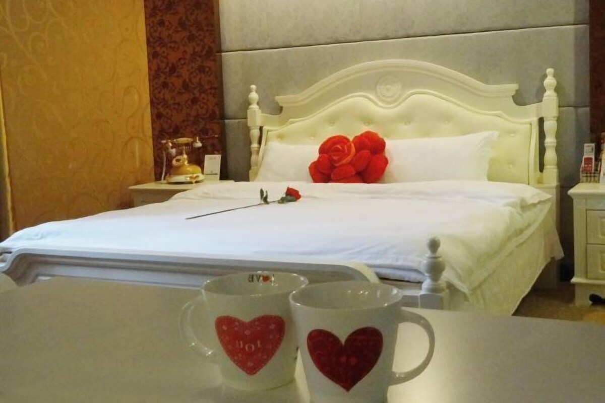A hotel room with a white bed, a nightstand with a telephone, and two heart-shaped mugs on the table.