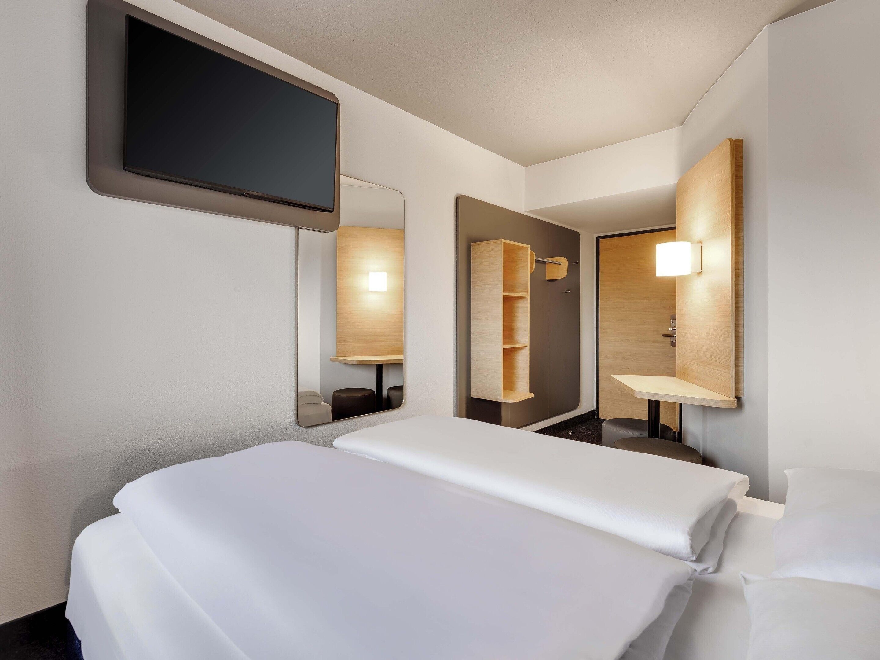 Una habitación de hotel con una cama, un televisor de pantalla plana, un espejo y una estantería de madera.