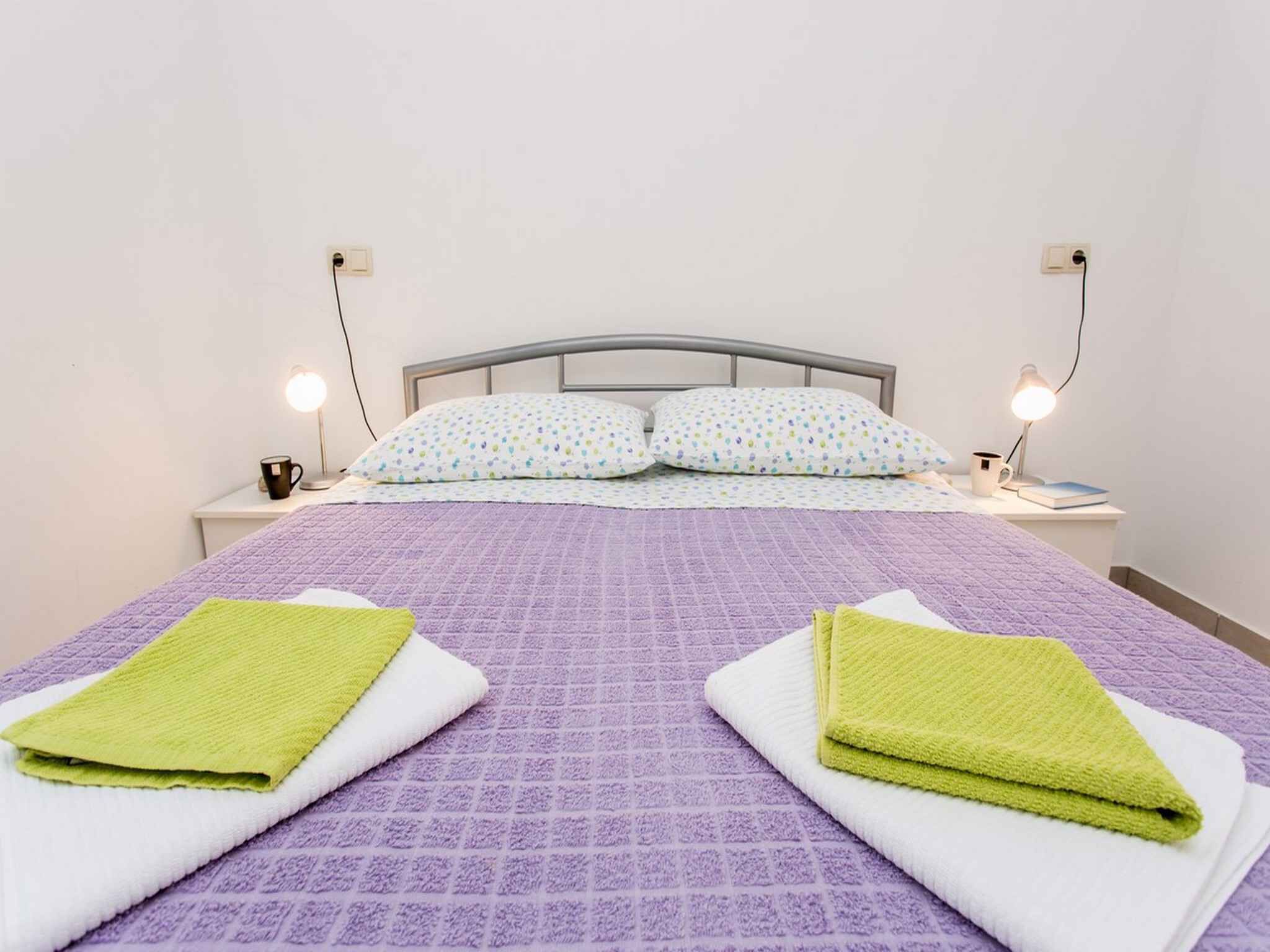A neatly made bed with a purple quilt, two green towels, and patterned pillows.