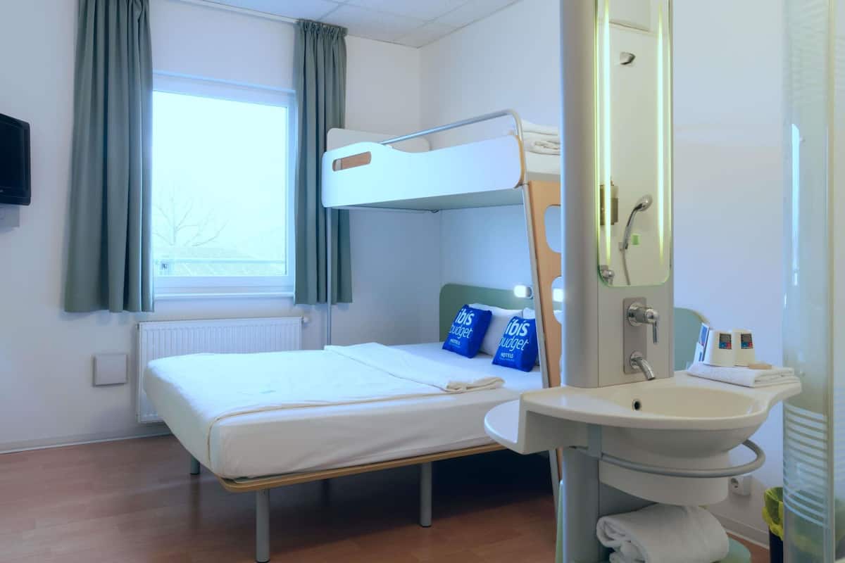 Ett hotellrum med en våningssäng, en enkelsäng, ett fönster med gardiner, ett element, ett handfat och en spegel.