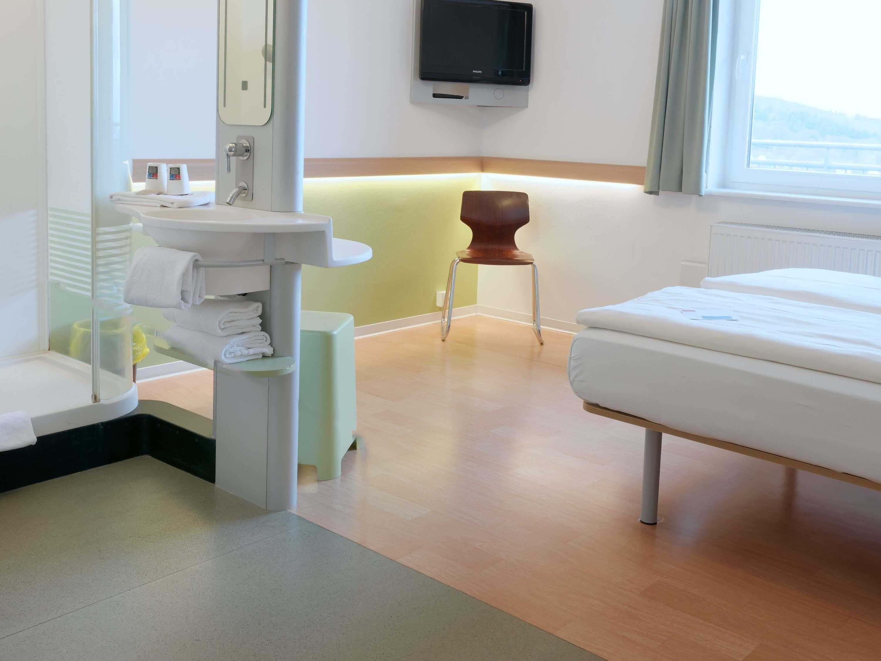 A hotel room with a bed, a bedside table, a small stool, a sink with a mirror, and a television mounted on the wall.