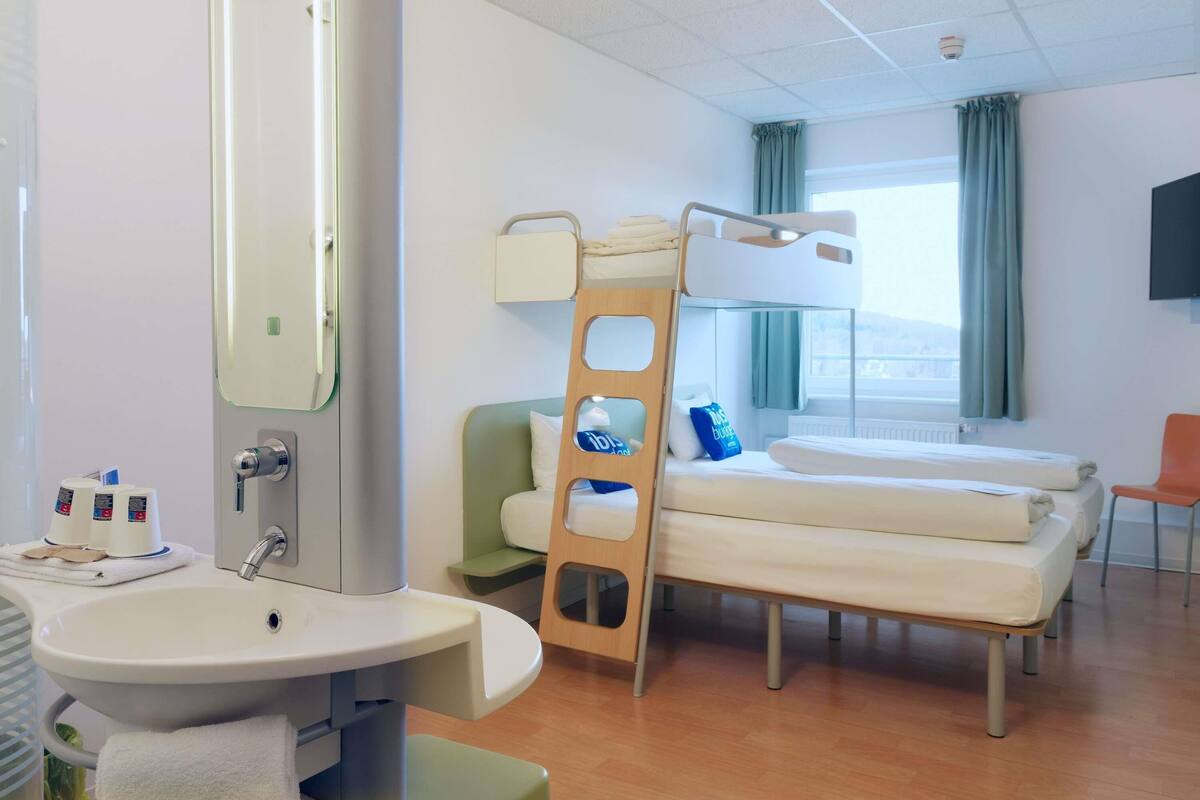 A hotel room with a bunk bed, a bedside table, a sink, a mirror, and a window with curtains.