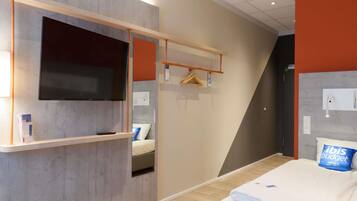 Habitación de hotel con cama, televisor montado en la pared, estante de madera y perchero.