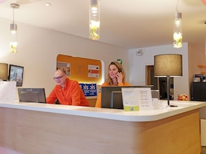 Un mostrador de recepción de hotel con dos empleados, un panel de señalización digital y una lámpara.