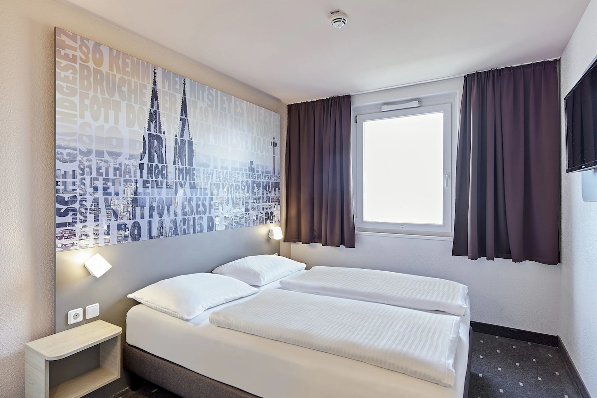 A hotel room with two beds, a large cityscape artwork on the wall, a window with curtains, and a small bedside table with a lamp.