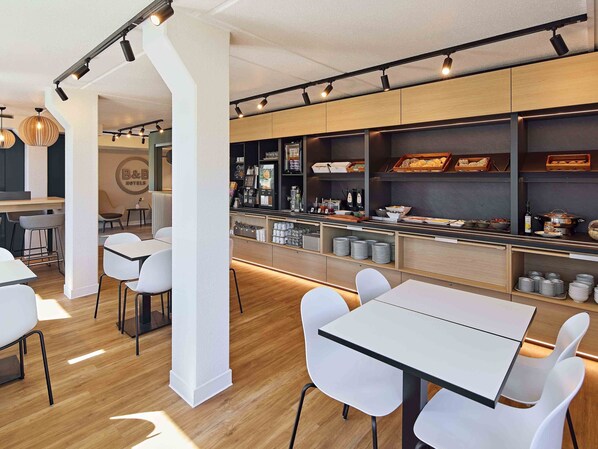 Un restaurant d’hôtel moderne avec des tables et des chaises blanches, un bar équipé d’une machine à café et des étagères remplies de produits alimentaires.
