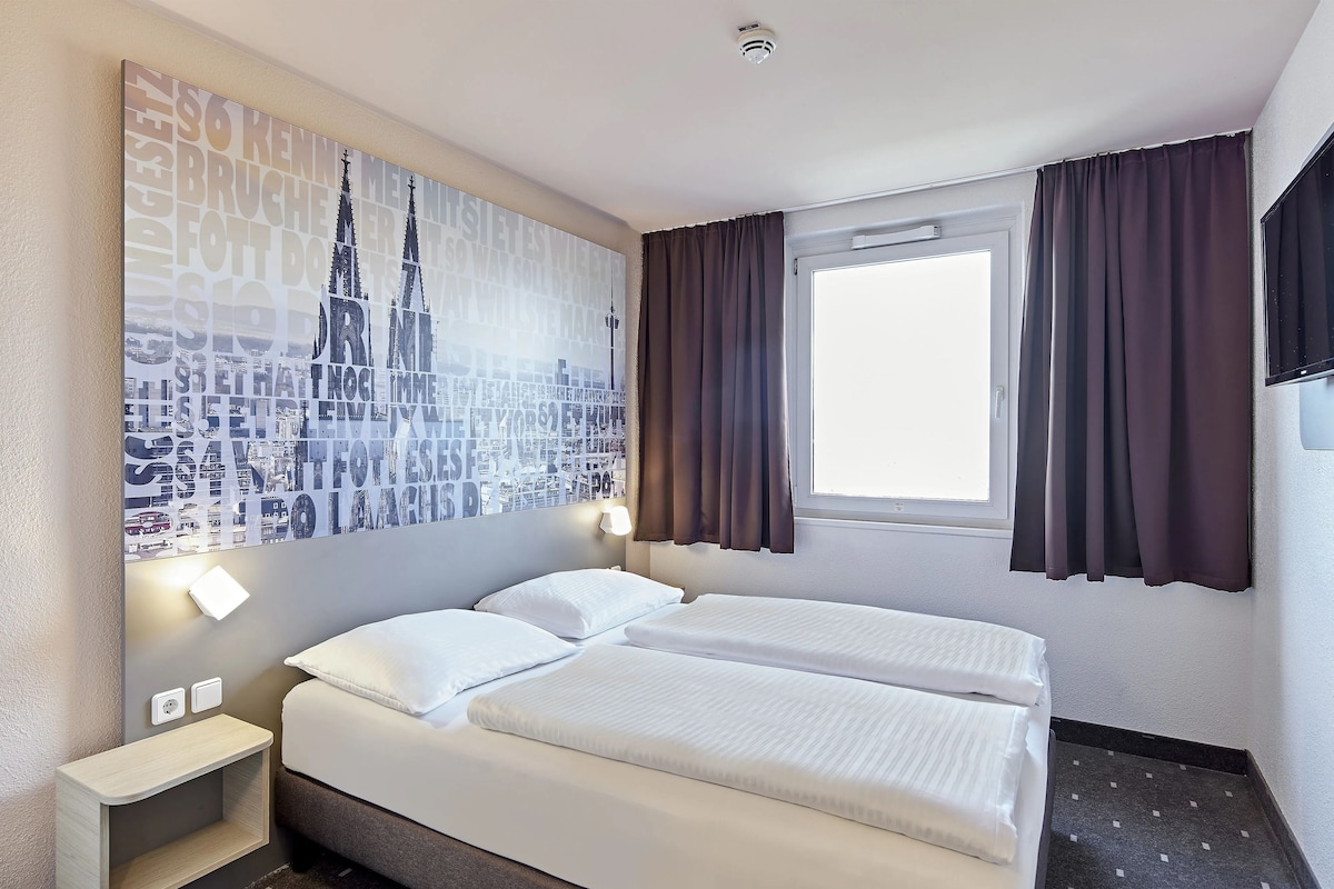 A hotel room with two beds, a large cityscape artwork on the wall, a window with curtains, and a small bedside table with a lamp.