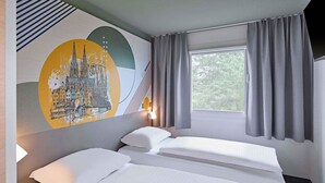 Ein Hotelzimmer mit zwei Betten, einem großen Fenster mit Vorhängen, einem Wandbild und einer Nachttischlampe.