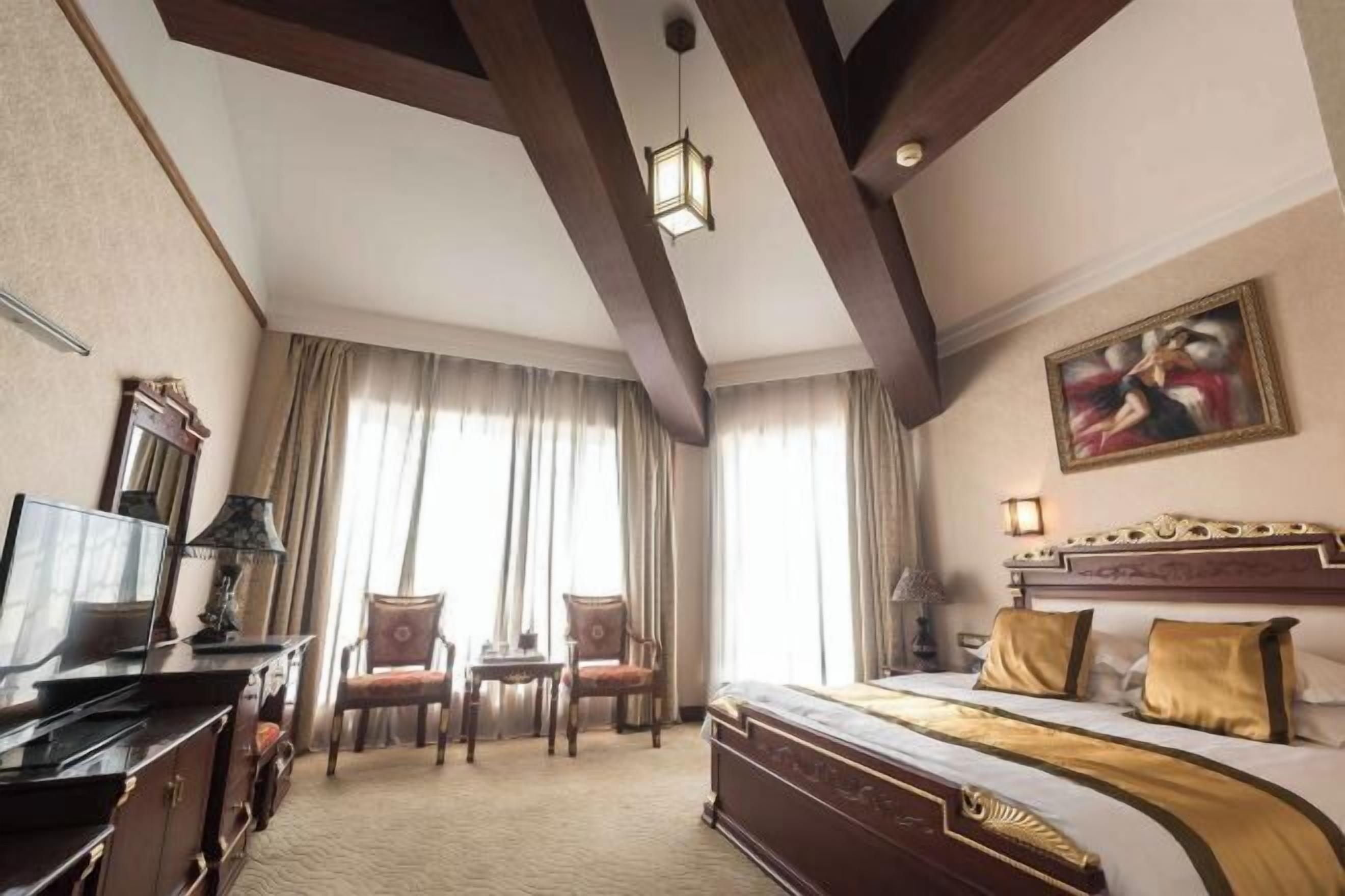 A hotel room with a large bed, a desk with a lamp, two chairs, a television, and a painting on the wall.