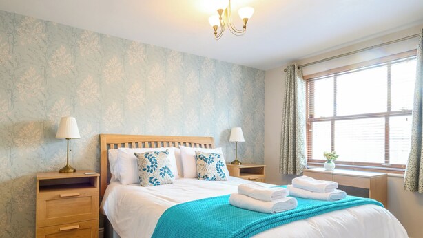 A neatly made bed with a blue blanket, two bedside tables with lamps, a window with curtains, and a wall with a floral pattern.