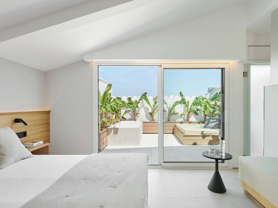 A modern hotel room with a large bed, a bedside table, a small table with a lamp, and a sliding glass door leading to a balcony with plants and a view of the sky.