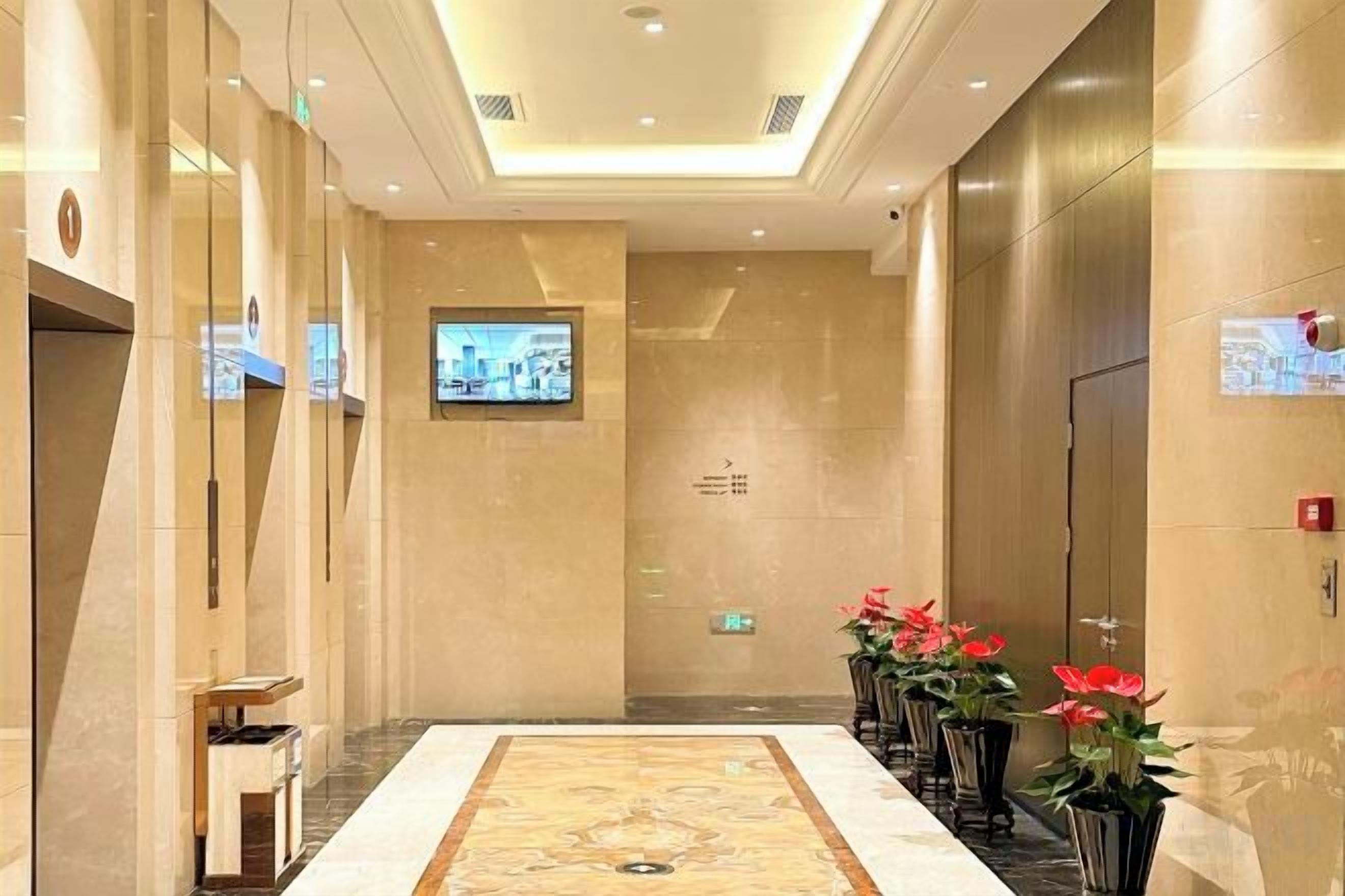 A hotel lobby with a marble floor, a reception desk, a television screen, and potted plants.
