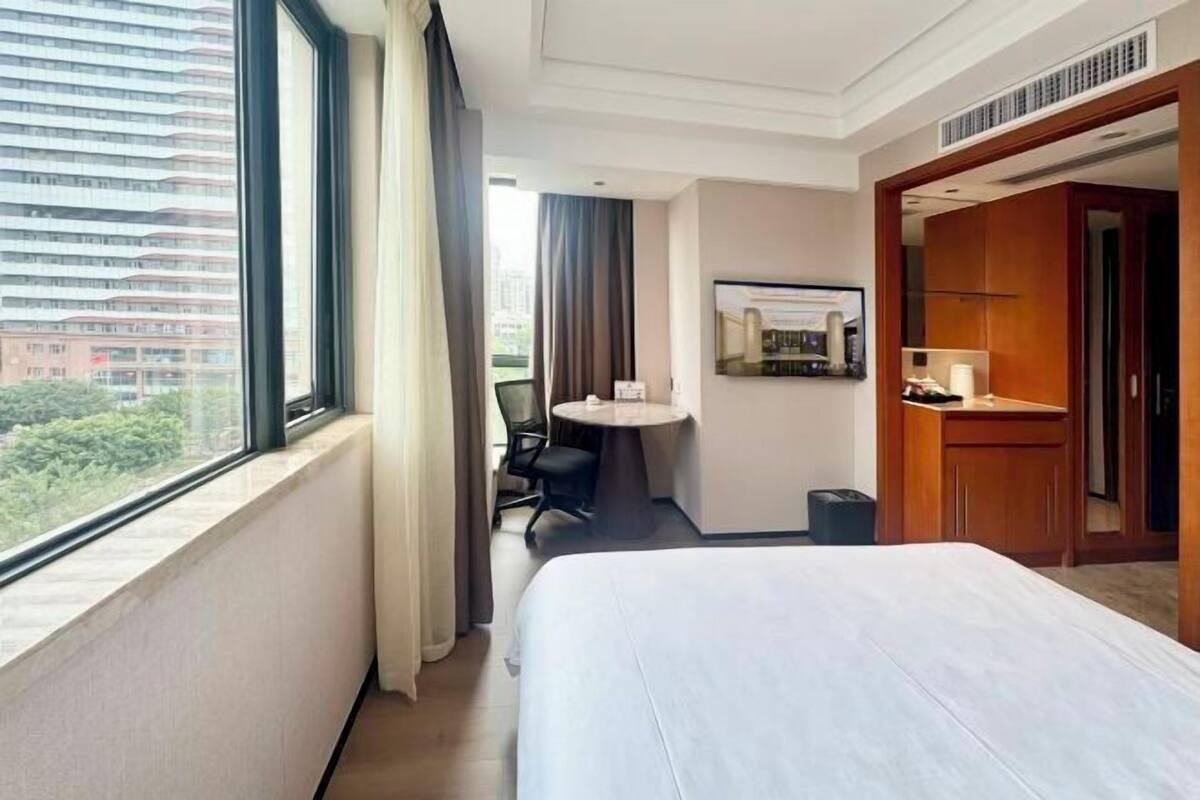 A hotel room with a bed, a desk, a chair, a small table, a trash bin, and a view of the city.