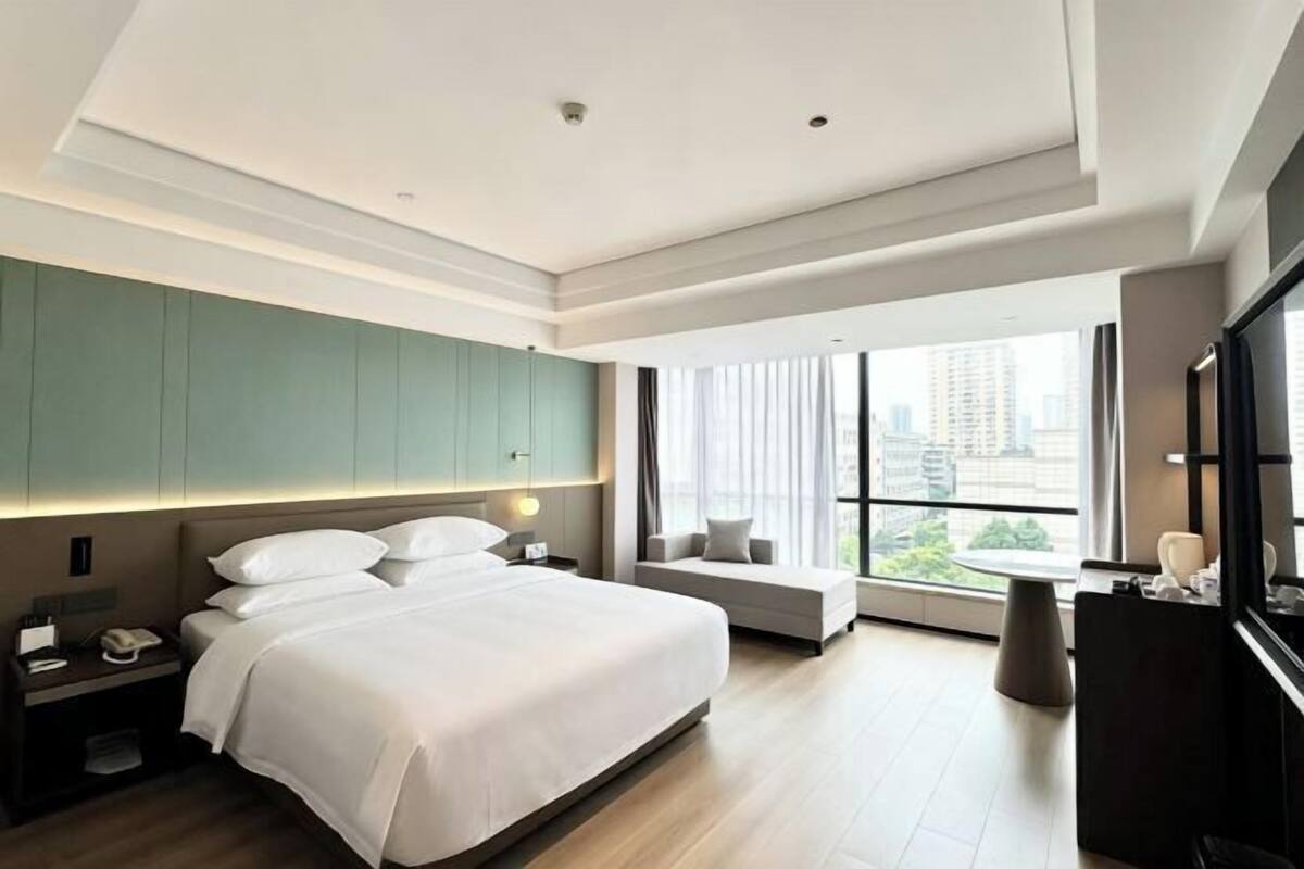 Una habitación de hotel moderna con una cama grande, un área de estar, un escritorio y un ventanal que ofrece vistas a la ciudad.