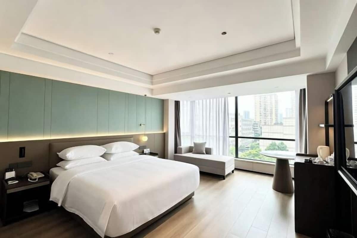 A modern hotel room with a large bed, a seating area, a desk, and a large window offering a city view.