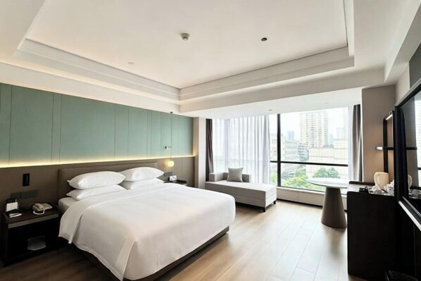 A modern hotel room with a large bed, a seating area, a desk, and a large window offering a city view.