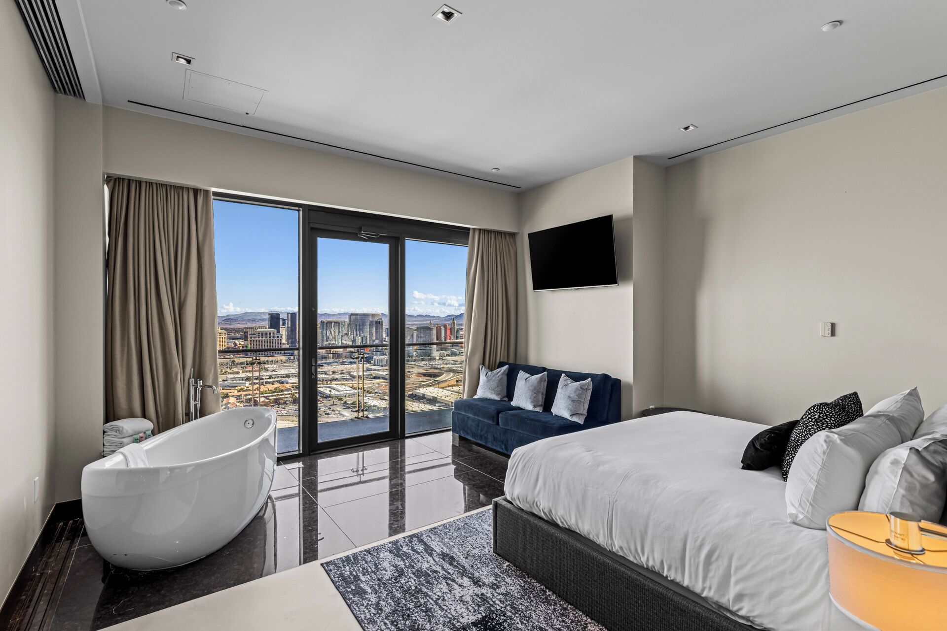 A modern hotel room with a large bed, a sofa, a bathtub, and a city view.