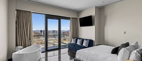 A modern hotel room with a large bed, a sofa, a bathtub, and a city view.