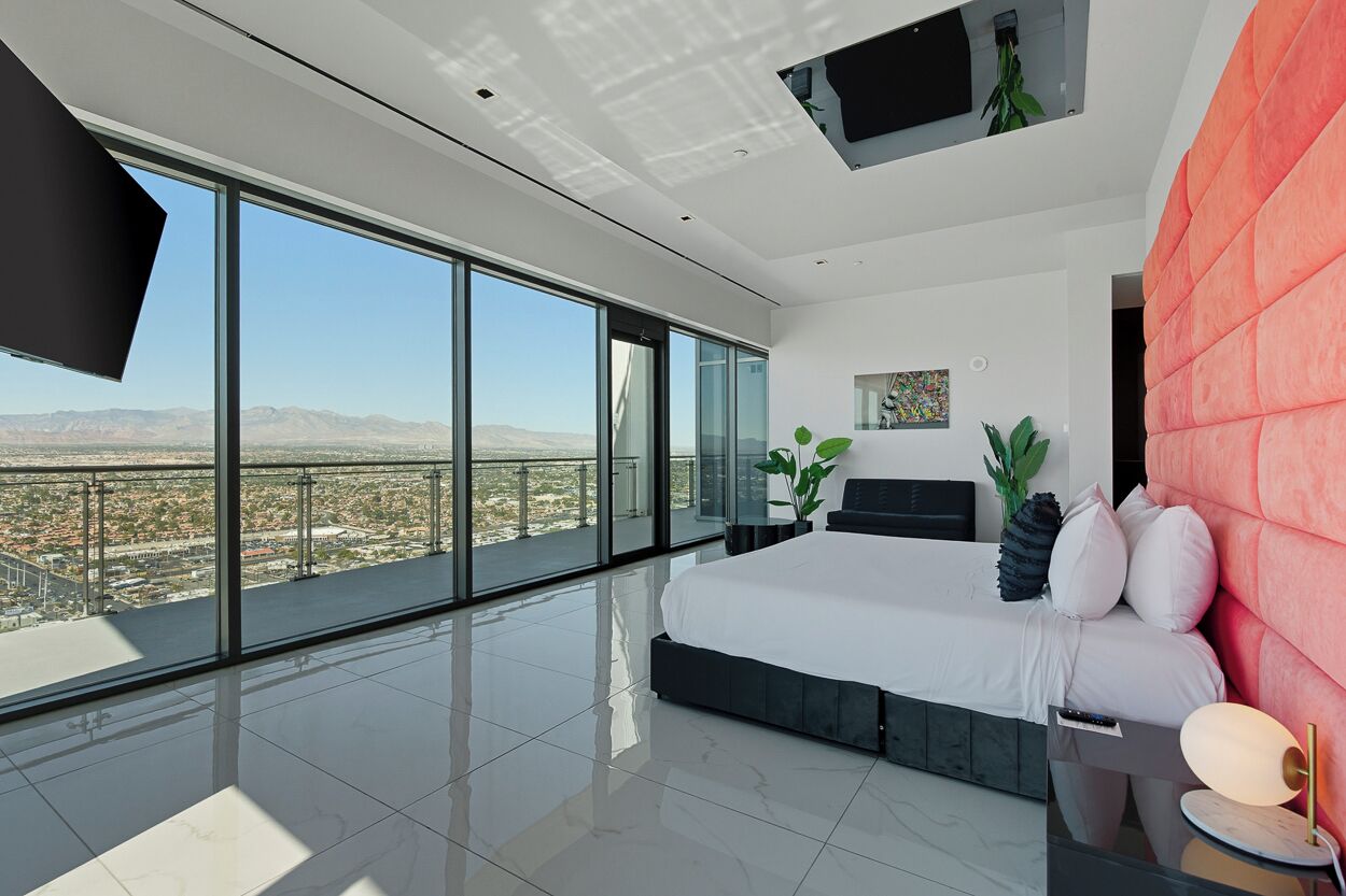 A modern bedroom with a large bed, a red accent wall, and a city view through floor-to-ceiling windows.