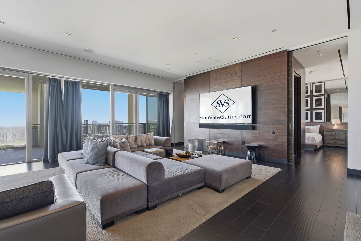 A modern living room with a large sectional sofa, a flat-screen TV mounted on the wall, and a city view.