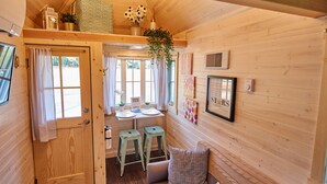 L’intérieur d’un chalet accueillant, avec une porte en bois, un petit coin repas avec des tabourets, un canapé avec des coussins et une plante suspendue.