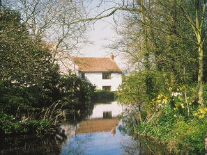 Et hvidt hus med et tegltag, omgivet af frodig vegetation og et roligt vandlegeme der spejler bygningen.