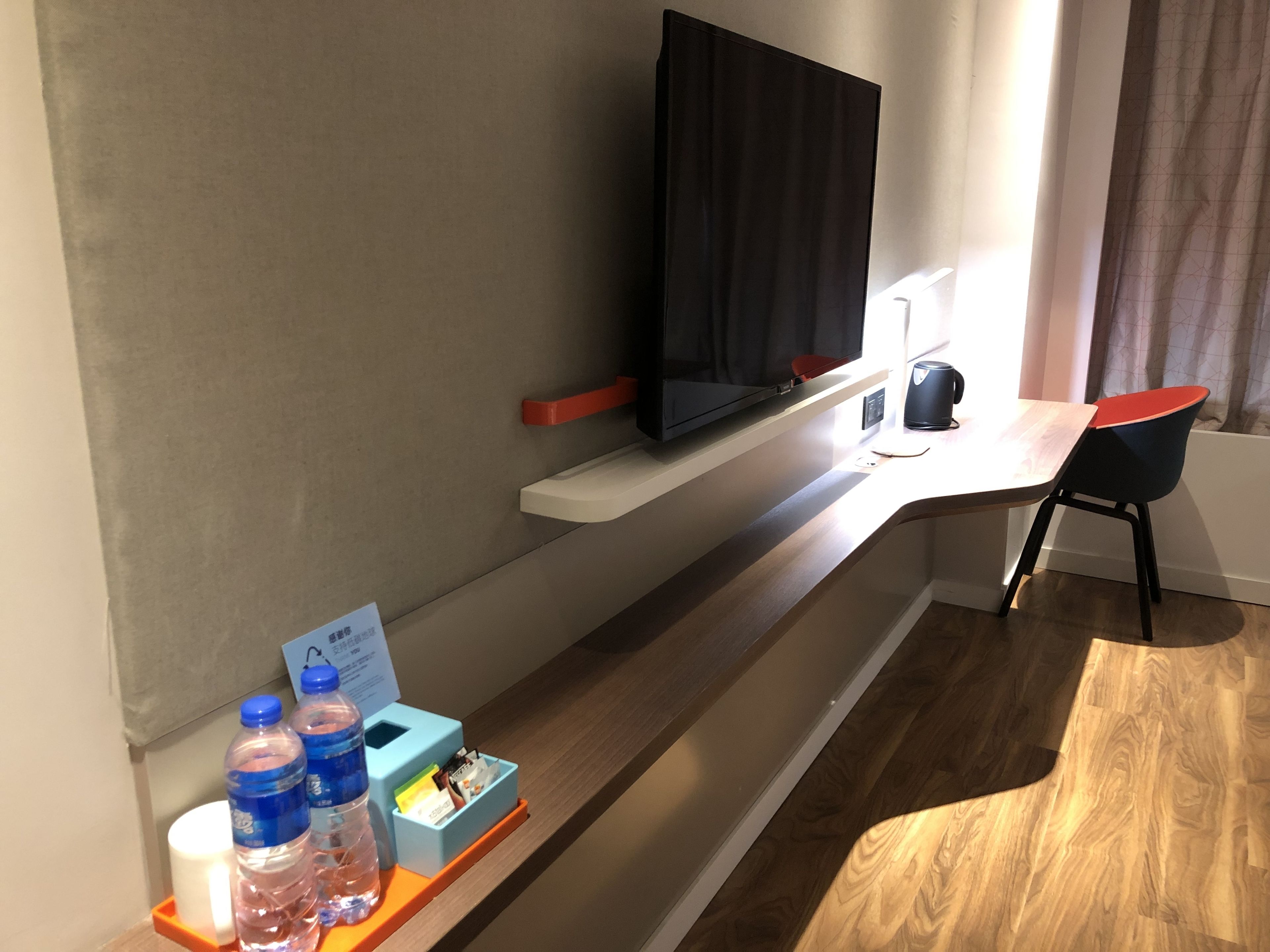 Camera d'albergo con TV a schermo piatto montata su uno scaffale bianco, una scrivania con una sedia, un dispenser d'acqua e una piccola scatola di fazzoletti.