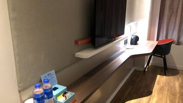 Habitación de hotel con televisión de pantalla plana montada en un estante blanco, un escritorio con una silla, dispensador de agua y una pequeña caja de pañuelos.