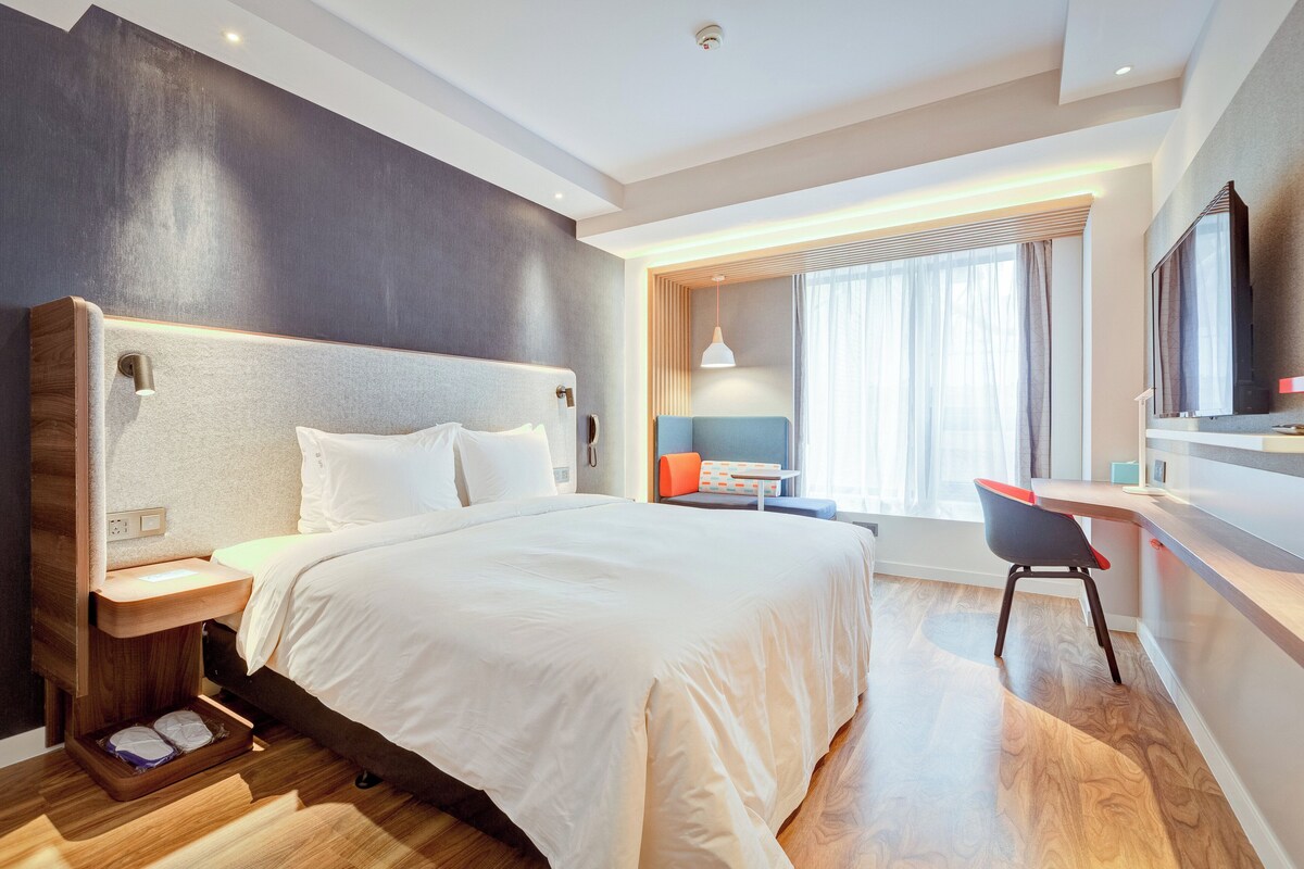 Ein modernes Hotelzimmer mit einem großen Bett, Nachttischen, einem Schreibtisch mit Stuhl und einem großen Fenster mit Vorhängen.