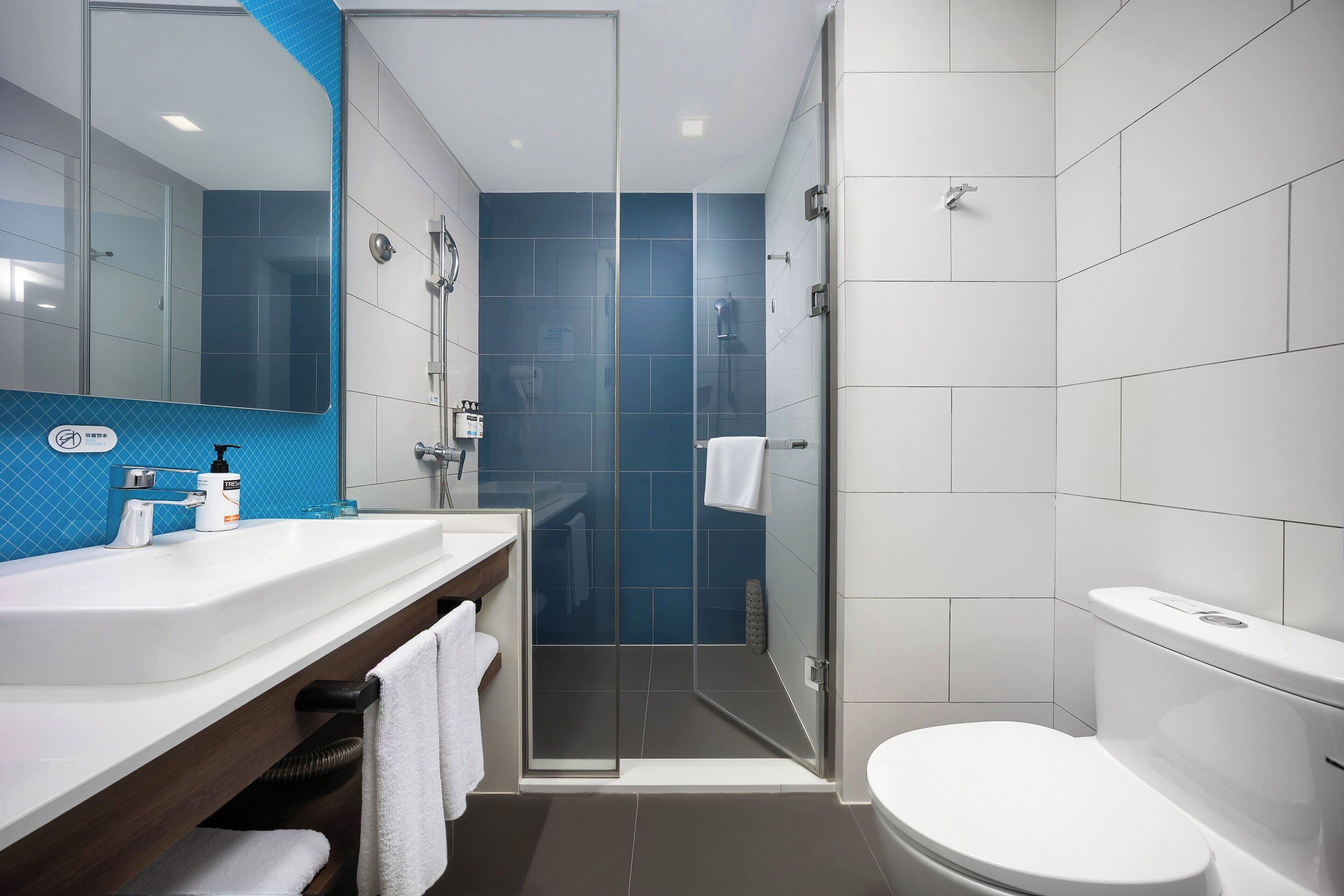 A modern bathroom with a white sink, mirror, and toilet, a glass-enclosed shower, and blue-tiled walls.