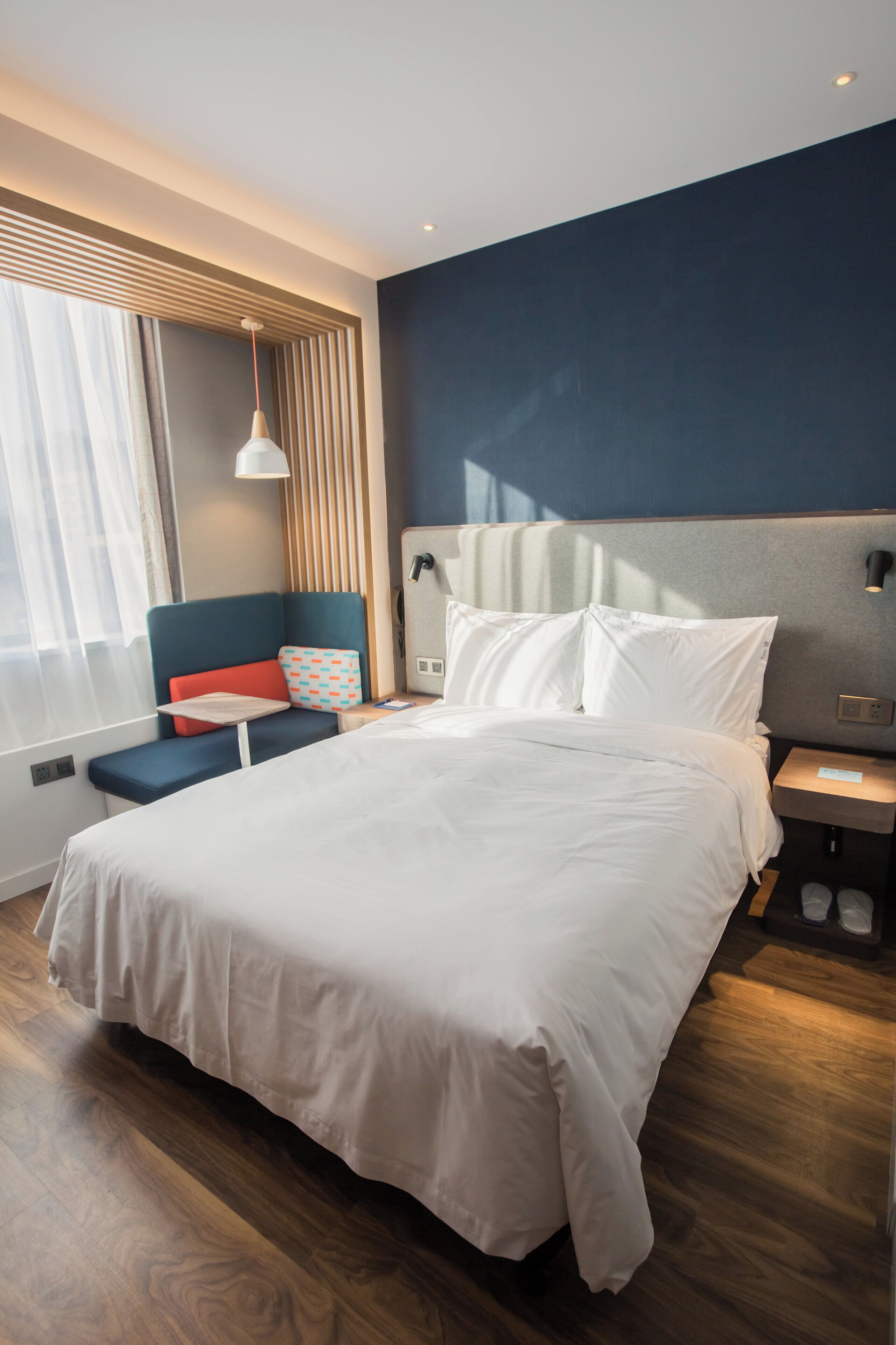 Ein modernes Hotelzimmer mit einem großen Bett, einem Nachttisch, einer Fenster mit Vorhängen und einer Wandlampe.