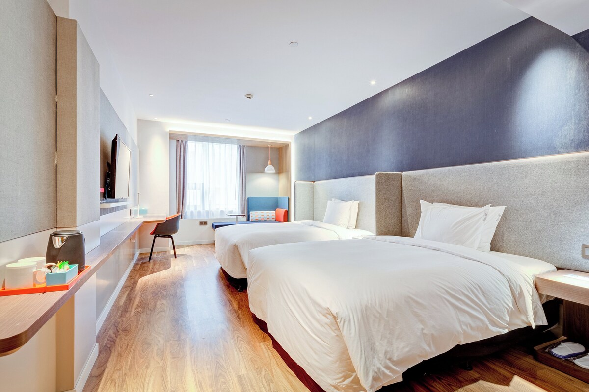 A hotel room with two beds, a desk, a chair, a coffee maker, and a window with curtains.