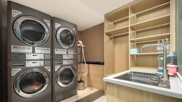 A laundry area with two stacked washing machines, a sink with a dish rack, and a wooden shelving unit.