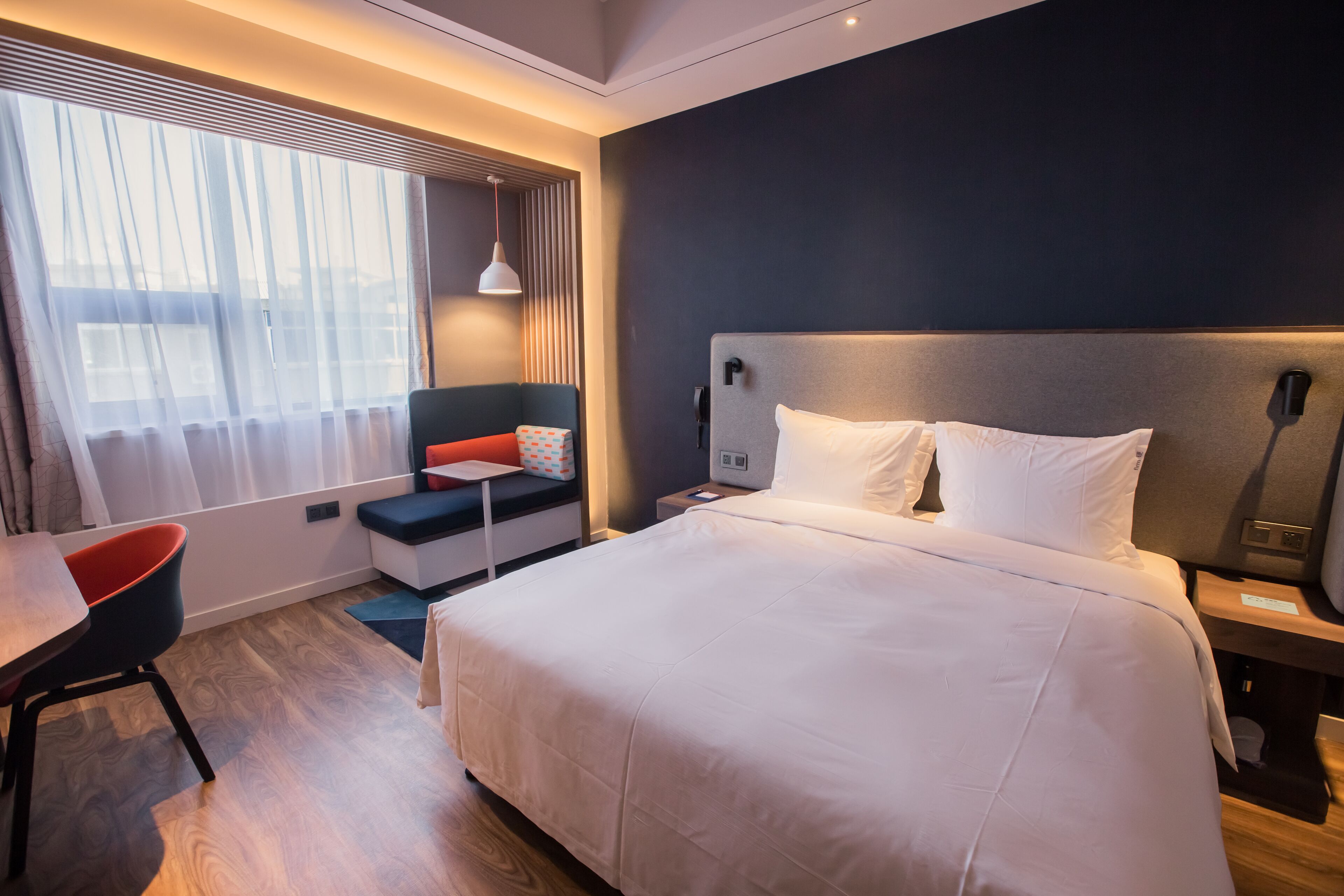 Ein modernes Hotelzimmer mit einem großen Bett, einem Schreibtisch, einem Stuhl und einem Fenster mit transparenten Vorhängen.