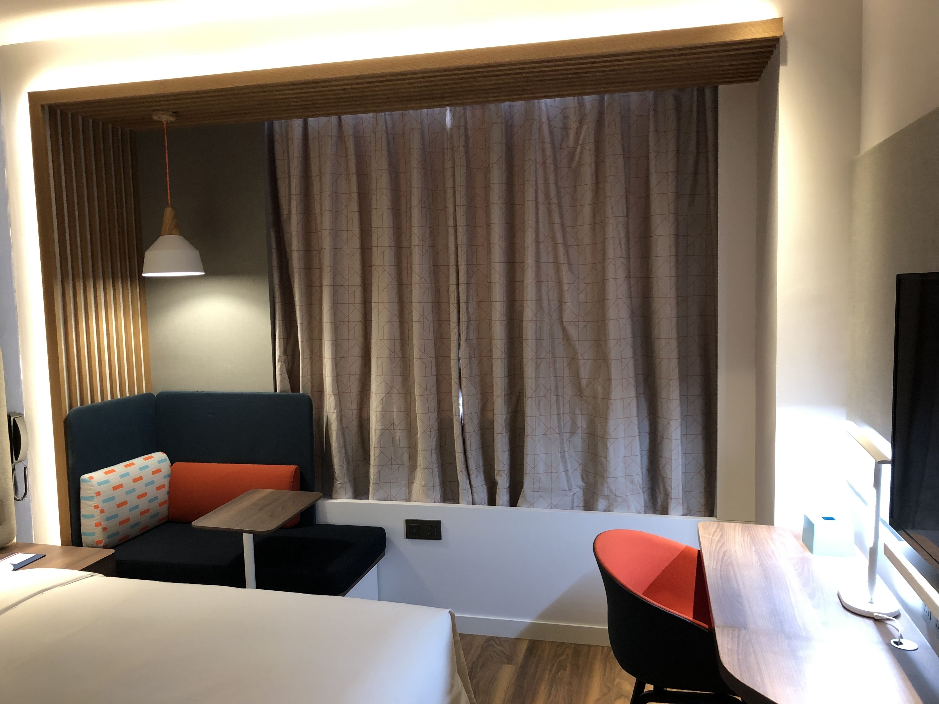 A modern hotel room with a bed, a desk, a chair, a sofa, and a large window with curtains.