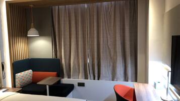 A modern hotel room with a bed, a desk, a chair, a sofa, and a large window with curtains.