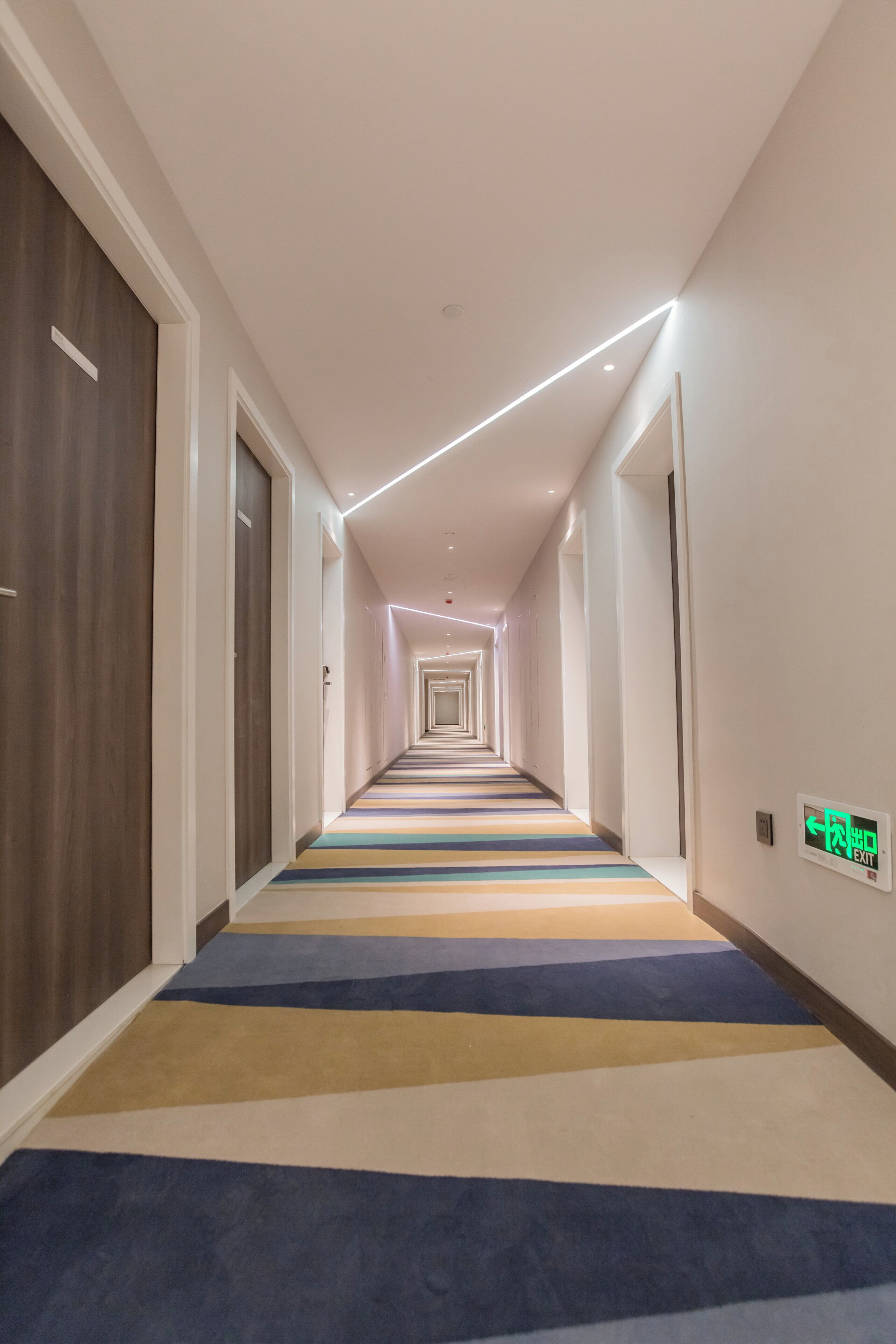 Un pasillo de hotel con alfombra a rayas, paredes blancas y una puerta de madera a la izquierda.