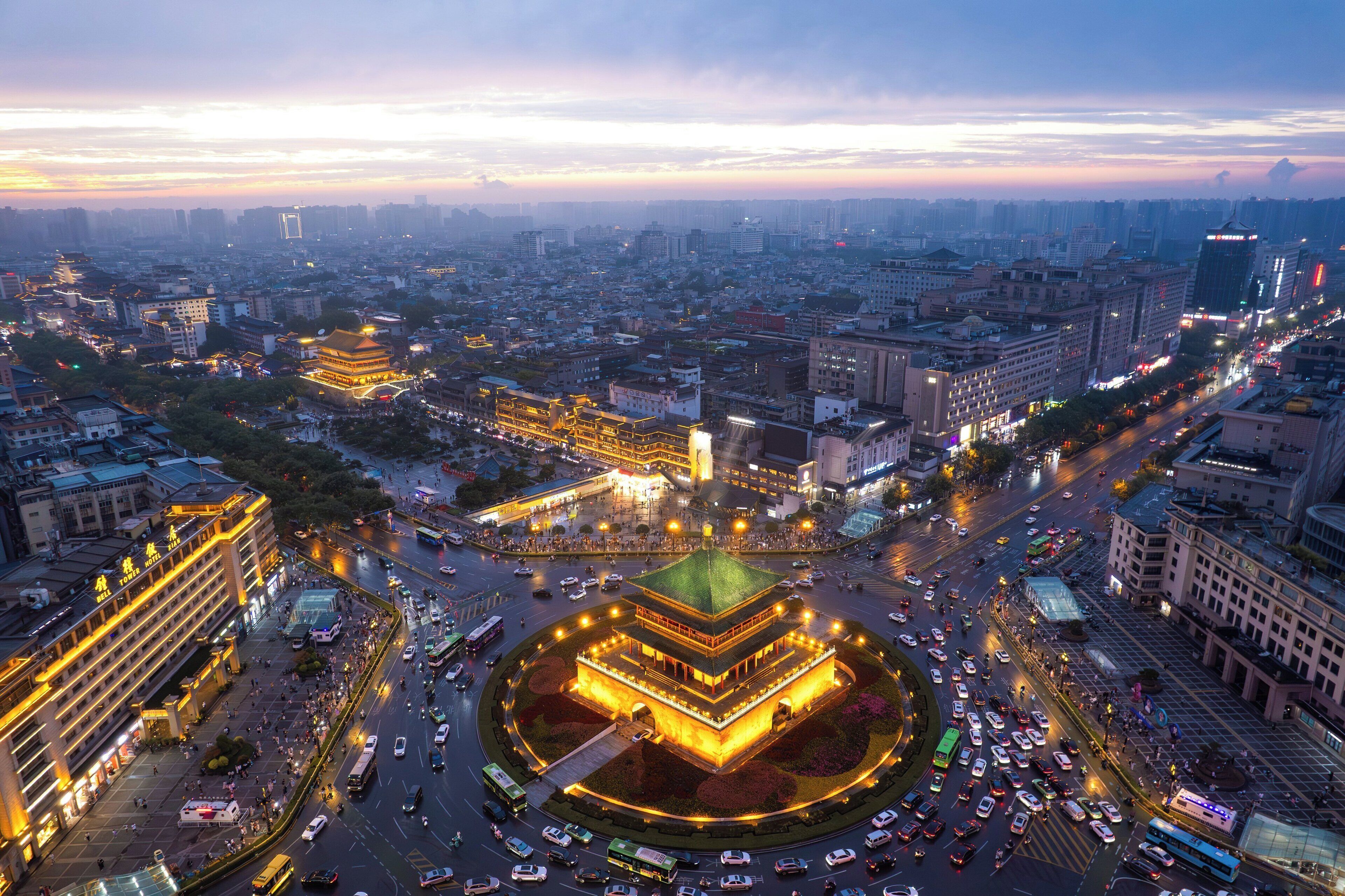 A cityscape with a prominent multi-story building featuring a central courtyard with a green roof, surrounded by a busy urban environment with various other buildings and streets.