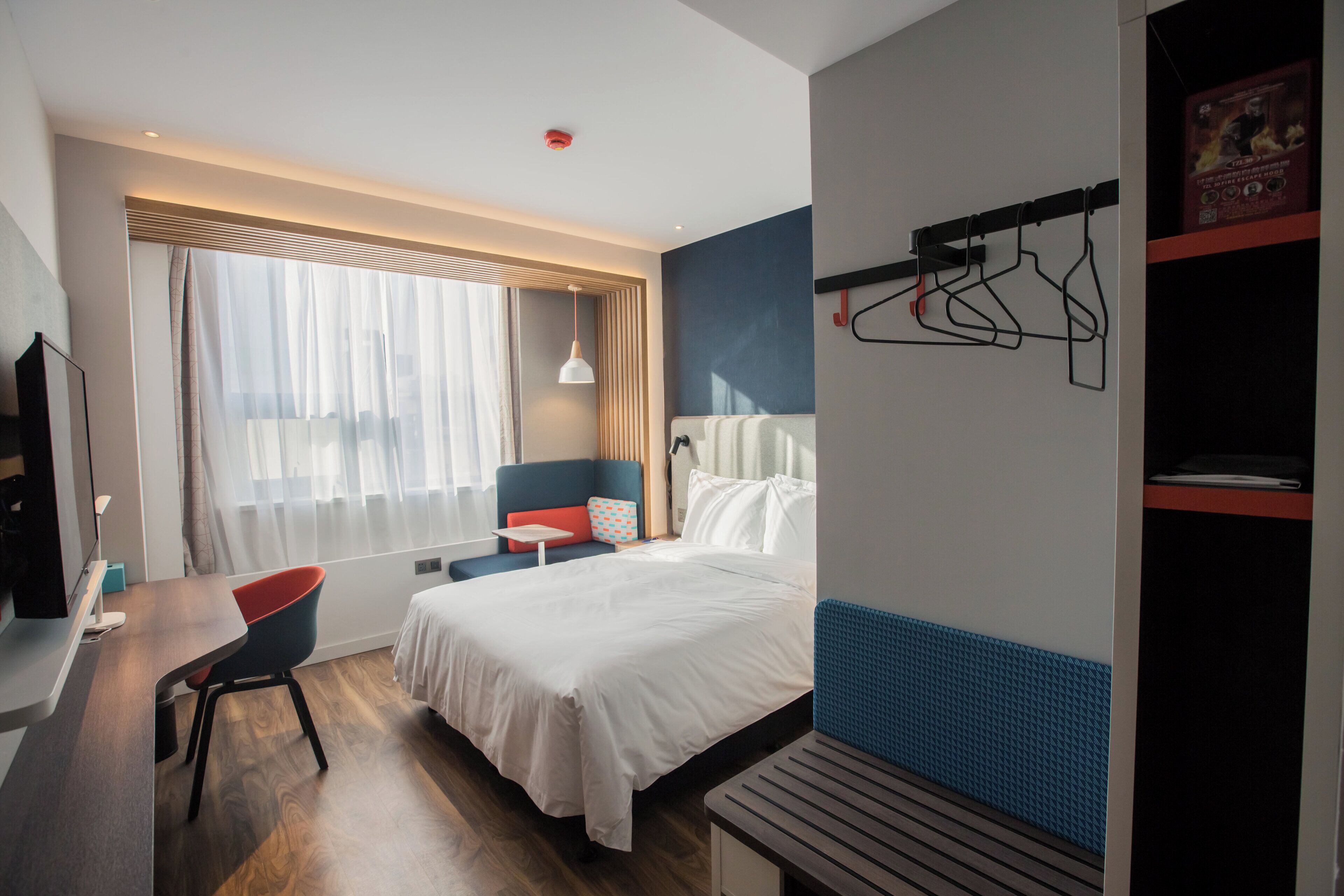 Una camera d'albergo con un letto, una scrivania, una sedia, una finestra con tende e un attaccapanni a parete.