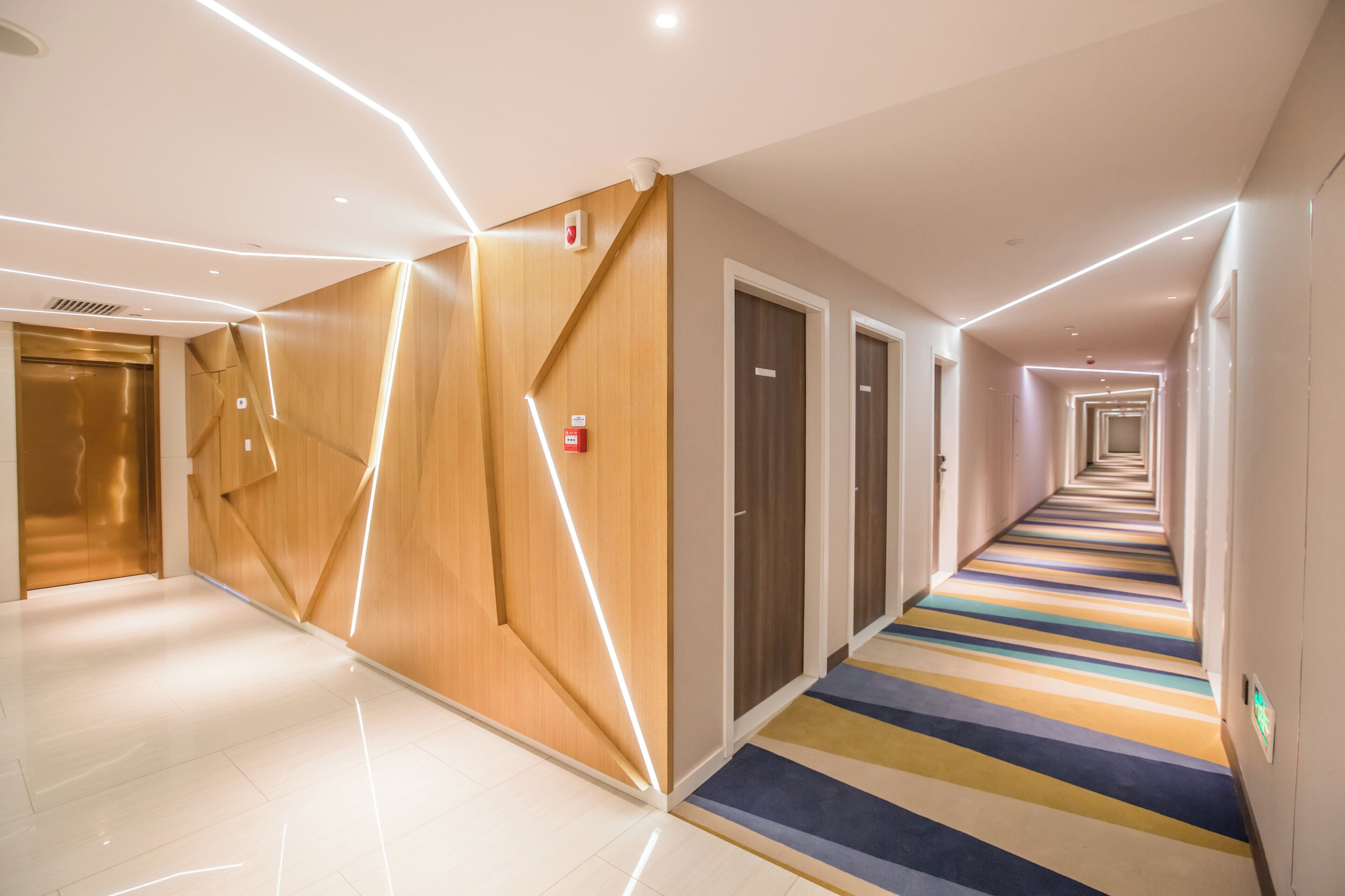 A modern hotel corridor with a reflective floor, wooden panel walls, and a striped carpet.