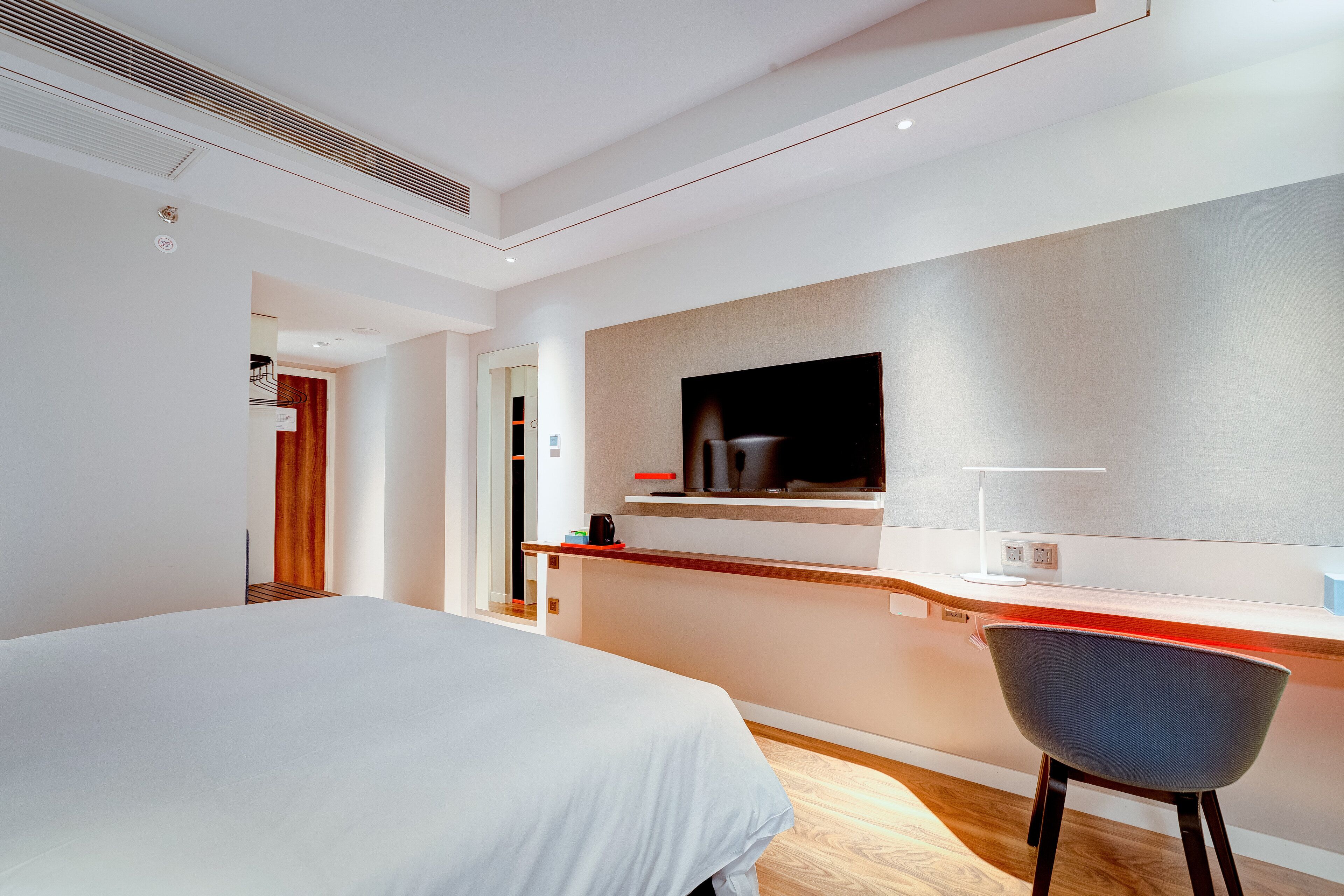Moderni hotellihuone, jossa on sänky, työpöytä tuolilla, seinälle asennettu taulutelevisio ja pieni keittonurkkaus.